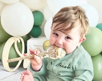 May include: A light green ribbed long-sleeve romper with the name Jacob embroidered in dark green thread on the chest. The garment features a button-up neckline and is worn by a child holding a wooden spoon covered in white cake frosting.