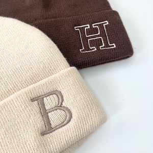 May include: Two knitted beanies, one brown with a white embroidered "H" and one cream with a brown embroidered "B".