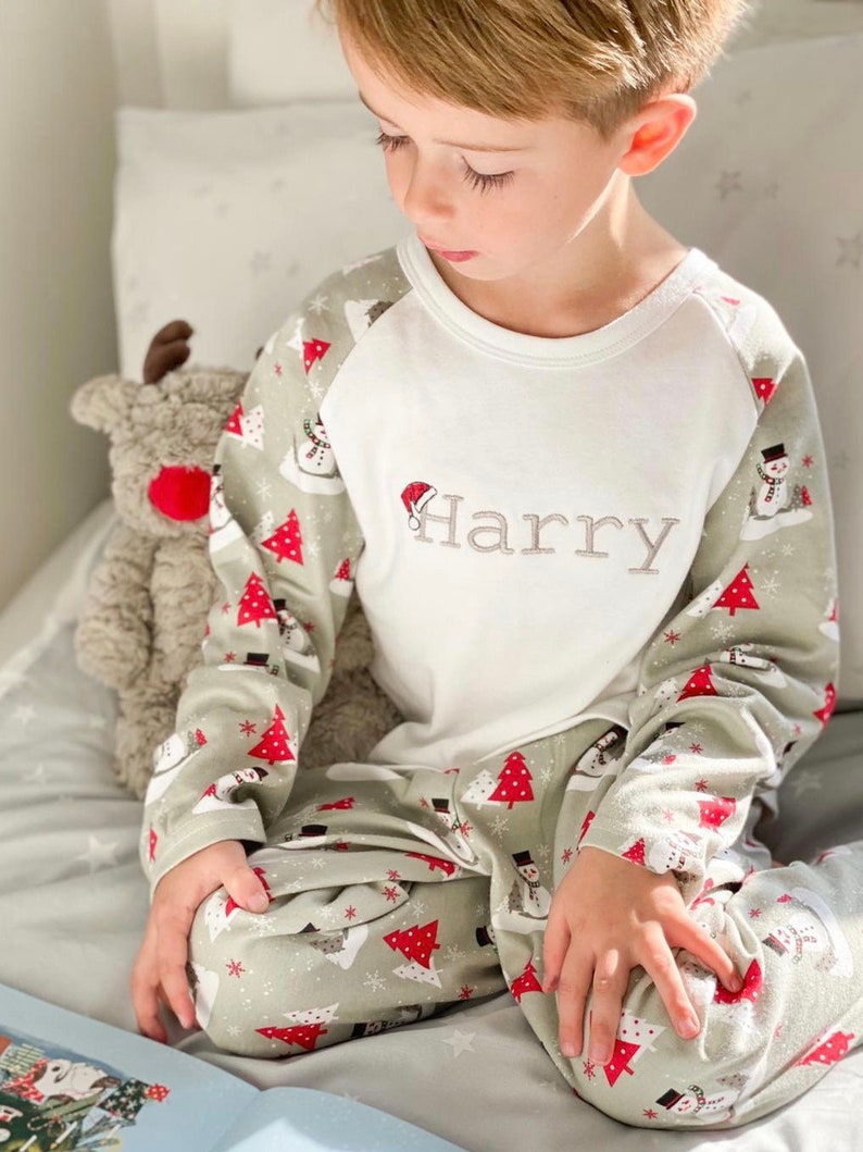 May include: A child wearing a white long-sleeved pajama top with the name "Harry" embroidered on the front. The pajama top has a green and white pattern with red Christmas trees and snowmen on the sleeves. The child is wearing matching green and white pajama pants with the same pattern.