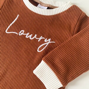 K&ouml;nnte beinhalten: Braunes Langarmshirt mit wei&szlig;em geripptem Besatz an Hals und &Auml;rmeln. Das Shirt hat den Namen "Loury" in wei&szlig; auf der Vorderseite gestickt.