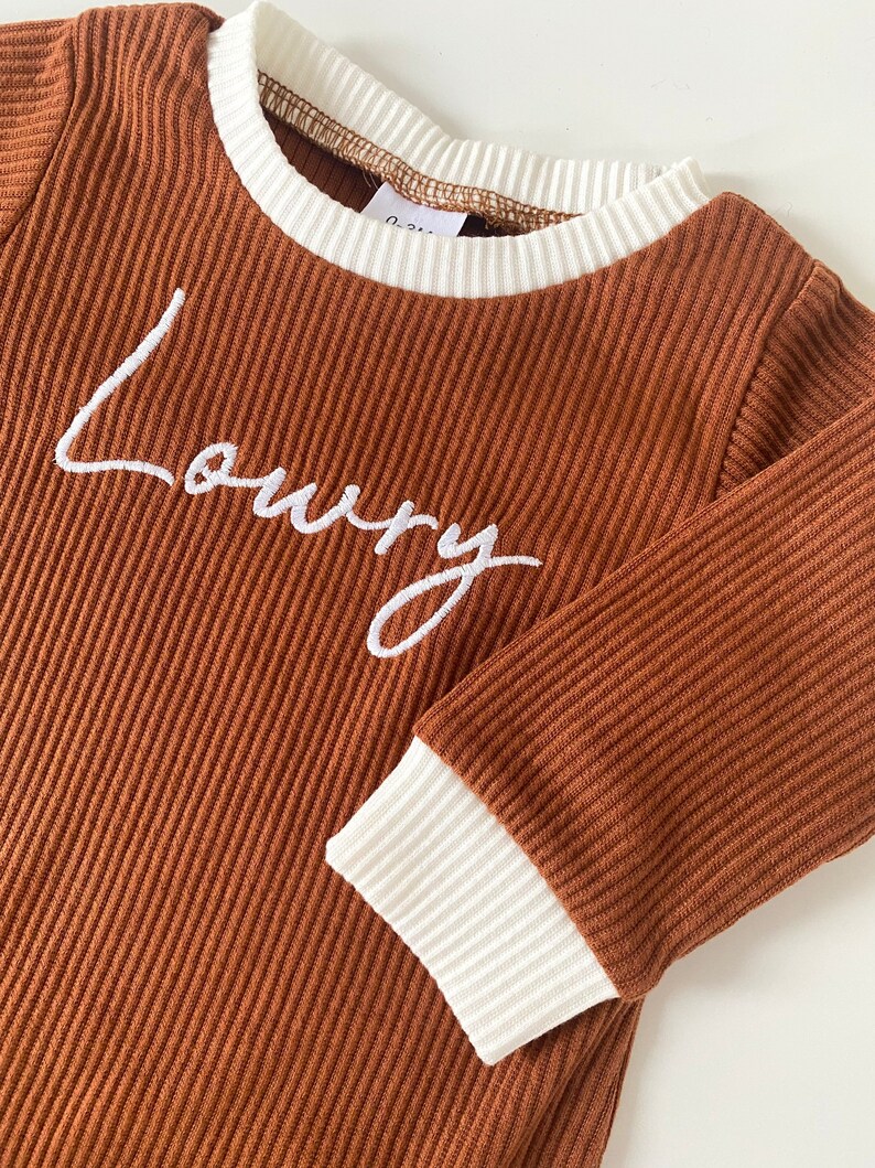 K&ouml;nnte beinhalten: Braunes Langarmshirt mit wei&szlig;em geripptem Besatz an Hals und &Auml;rmeln. Das Shirt hat den Namen "Loury" in wei&szlig; auf der Vorderseite gestickt.