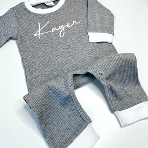 K&ouml;nnte beinhalten: Grauer Langarm-Baby-Strampler mit wei&szlig;en gerippten Abschl&uuml;ssen und wei&szlig;em gesticktem Namen "Kayen".
