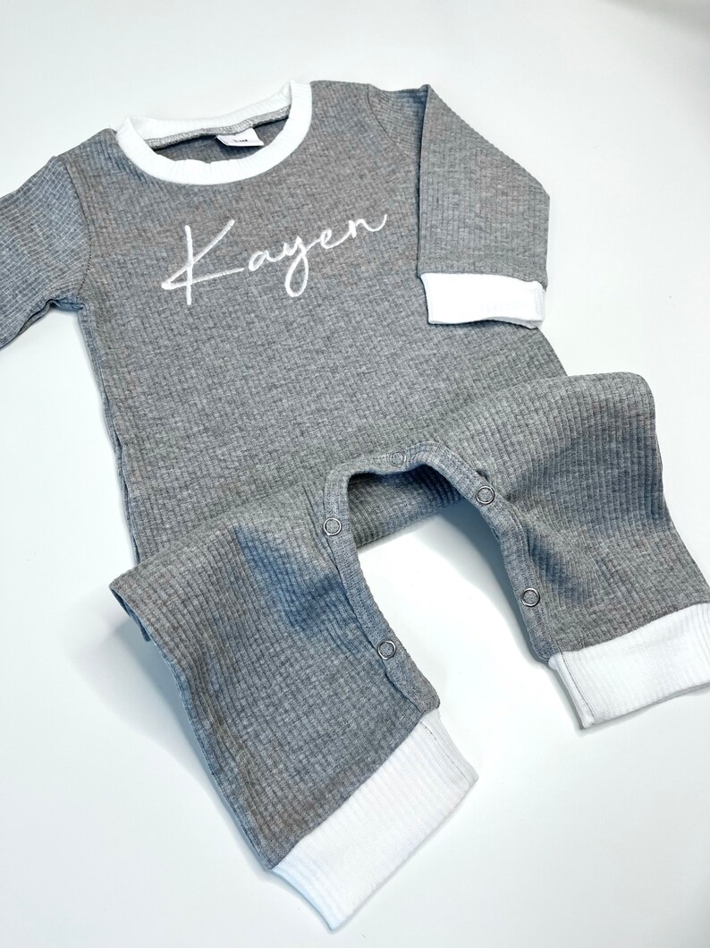 K&ouml;nnte beinhalten: Grauer Langarm-Baby-Strampler mit wei&szlig;en gerippten Abschl&uuml;ssen und wei&szlig;em gesticktem Namen "Kayen".