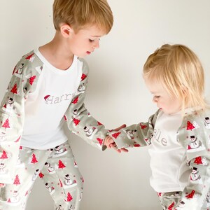 May include: Two children wearing matching green and white pajamas with a snowman pattern. The pajamas have long sleeves and long pants. The boy's pajamas have the name "Harry" embroidered on the front. The girl's pajamas have the name "Ellie" embroidered on the front.