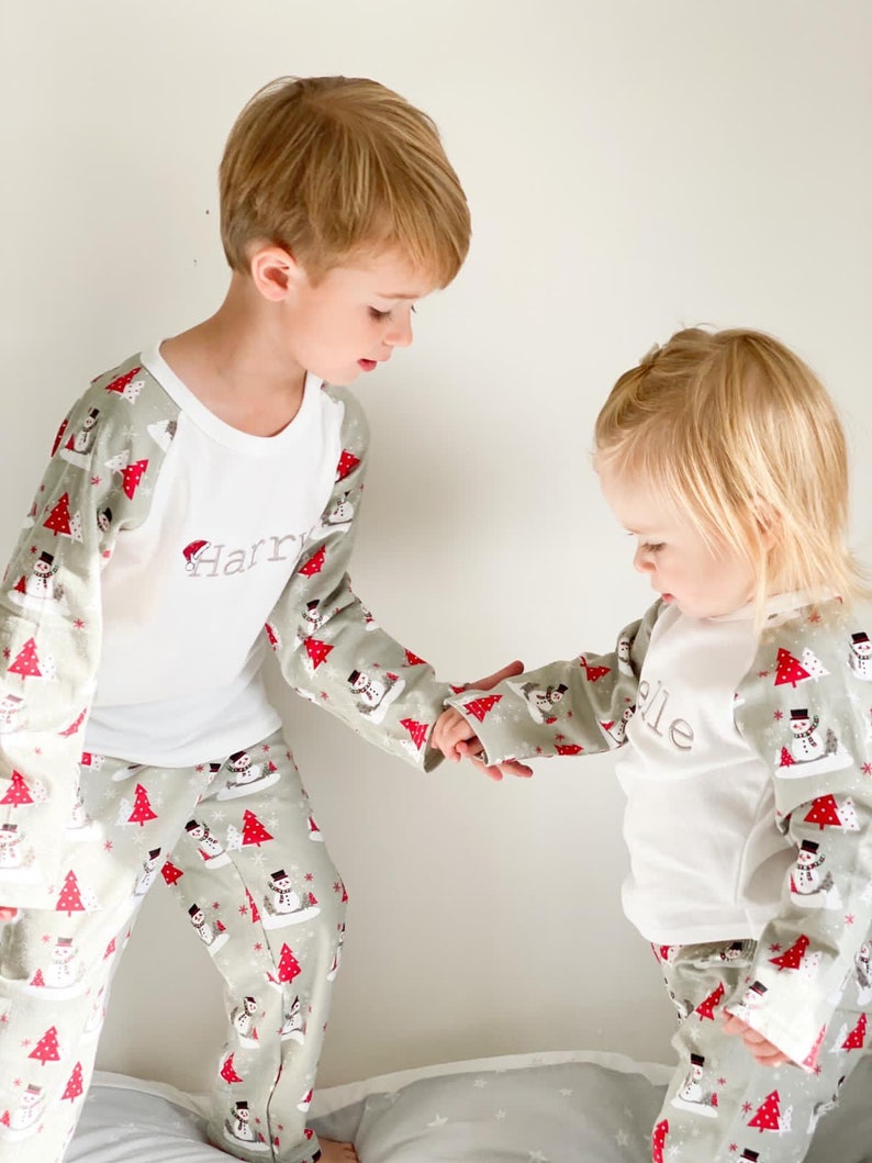 May include: Two children wearing matching green and white pajamas with a snowman pattern. The pajamas have long sleeves and long pants. The boy's pajamas have the name "Harry" embroidered on the front. The girl's pajamas have the name "Ellie" embroidered on the front.