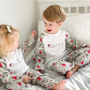 May include: Two children wearing matching green and white pajamas with a snowman and Christmas tree pattern. The pajamas have the names "Belle" and "Harry" embroidered on the front. The children are sitting on a bed and smiling at each other.