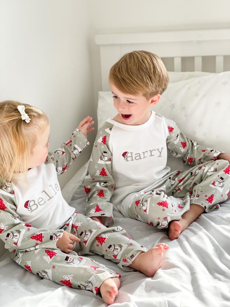 May include: Two children wearing matching green and white pajamas with a snowman and Christmas tree pattern. The pajamas have the names "Belle" and "Harry" embroidered on the front. The children are sitting on a bed and smiling at each other.