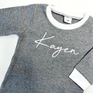 K&ouml;nnte beinhalten: Graues Langarmshirt mit wei&szlig;em geripptem Saum an Ausschnitt und &Auml;rmelb&uuml;ndchen. Das Shirt tr&auml;gt den Namen "Kayen" in wei&szlig;er Kursivschrift auf der Vorderseite.