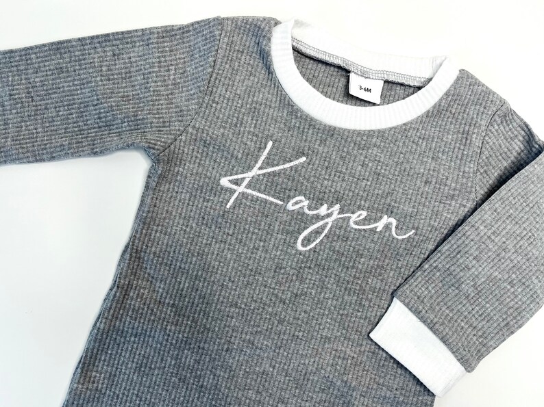 K&ouml;nnte beinhalten: Graues Langarmshirt mit wei&szlig;em geripptem Saum an Ausschnitt und &Auml;rmelb&uuml;ndchen. Das Shirt tr&auml;gt den Namen "Kayen" in wei&szlig;er Kursivschrift auf der Vorderseite.