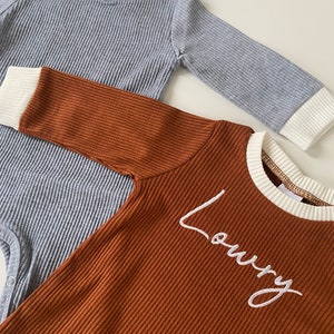 K&ouml;nnte beinhalten: Zwei lang&auml;rmelige Baby-Bodys. Einer ist grau mit wei&szlig;em Besatz und der andere ist braun mit wei&szlig;em Besatz und dem Namen "Loury" auf der Vorderseite gestickt.