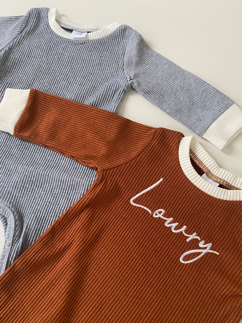 K&ouml;nnte beinhalten: Zwei lang&auml;rmelige Baby-Bodys. Einer ist grau mit wei&szlig;em Besatz und der andere ist braun mit wei&szlig;em Besatz und dem Namen "Loury" auf der Vorderseite gestickt.