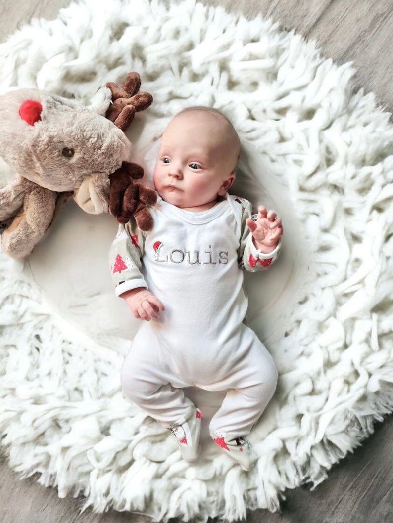 May include: A baby wearing a white onesie with red and green trim and the name "Louis" embroidered on the front. The baby is lying on a white fluffy rug with a brown reindeer stuffed animal.