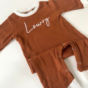 K&ouml;nnte beinhalten: Ein brauner gerippter lang&auml;rmliger Baby-Strampler mit wei&szlig;en B&uuml;ndchen und dem Namen "Lowry" auf der Vorderseite gestickt.