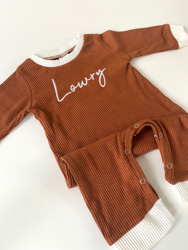 K&ouml;nnte beinhalten: Ein brauner gerippter lang&auml;rmliger Baby-Strampler mit wei&szlig;en B&uuml;ndchen und dem Namen "Lowry" auf der Vorderseite gestickt.