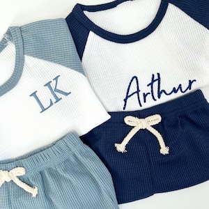 Könnte beinhalten: Zwei Waffelstrick-Baby-Outfits, eines blau und eines marineblau, mit weißen Waffelstrick-Langarmshirts. Das blaue Outfit hat den Namen "LK" auf das Shirt gestickt und das marineblaue Outfit hat den Namen "Arthur" auf das Shirt gestickt.