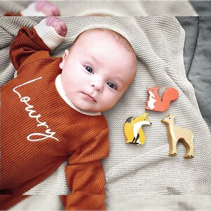 K&ouml;nnte beinhalten: Ein Baby tr&auml;gt einen lang&auml;rmeligen Strampler in Rostfarbe mit dem Wort "Lovey" bestickt auf der Vorderseite. Das Baby liegt auf einer wei&szlig;en Strickdecke mit drei Holztieren: einem Fuchs, einem Eichh&ouml;rnchen und einem Reh.