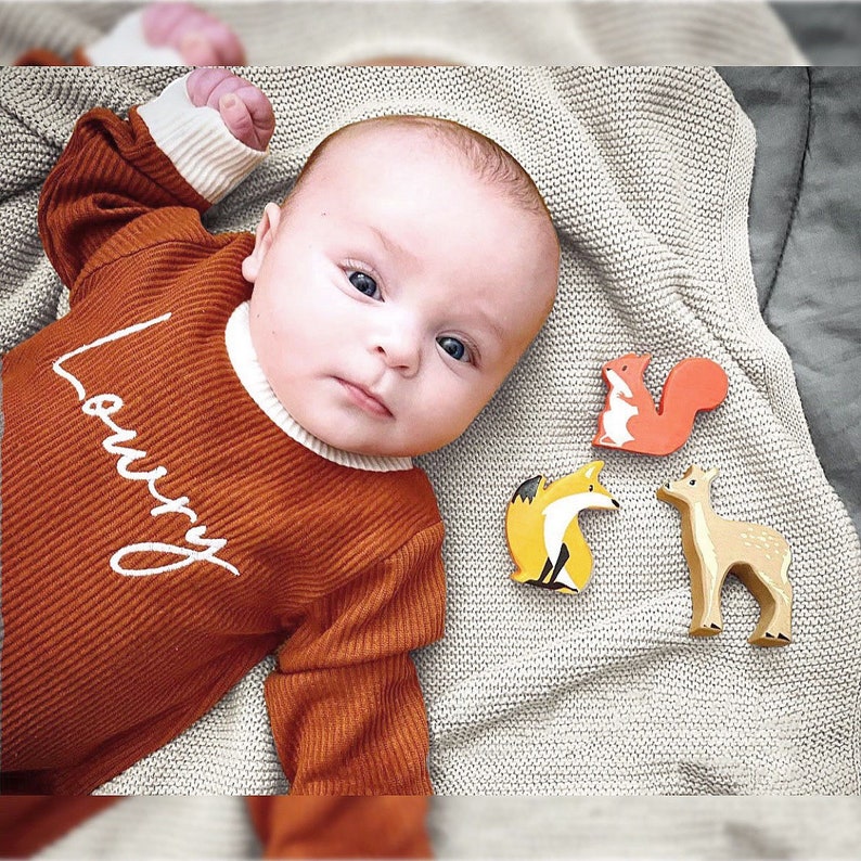 K&ouml;nnte beinhalten: Ein Baby tr&auml;gt einen lang&auml;rmeligen Strampler in Rostfarbe mit dem Wort "Lovey" bestickt auf der Vorderseite. Das Baby liegt auf einer wei&szlig;en Strickdecke mit drei Holztieren: einem Fuchs, einem Eichh&ouml;rnchen und einem Reh.