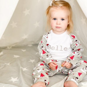 May include: A young child wearing a white long-sleeved shirt with the name "Belle" embroidered on the front. The child is wearing grey and white pajamas with a snowman and Christmas tree pattern. The child is sitting inside a teepee with a white star pattern.