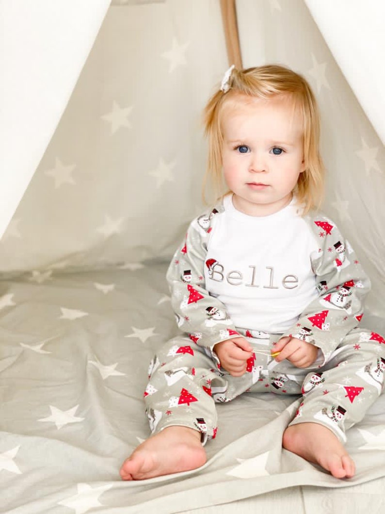 May include: A young child wearing a white long-sleeved shirt with the name "Belle" embroidered on the front. The child is wearing grey and white pajamas with a snowman and Christmas tree pattern. The child is sitting inside a teepee with a white star pattern.