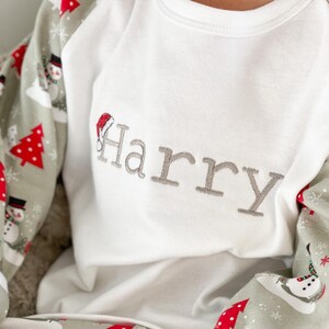 May include: White long-sleeve pajamas with a green and red snowman pattern. The top has a white collar and a gray embroidered name "Harry" with a red Santa hat.