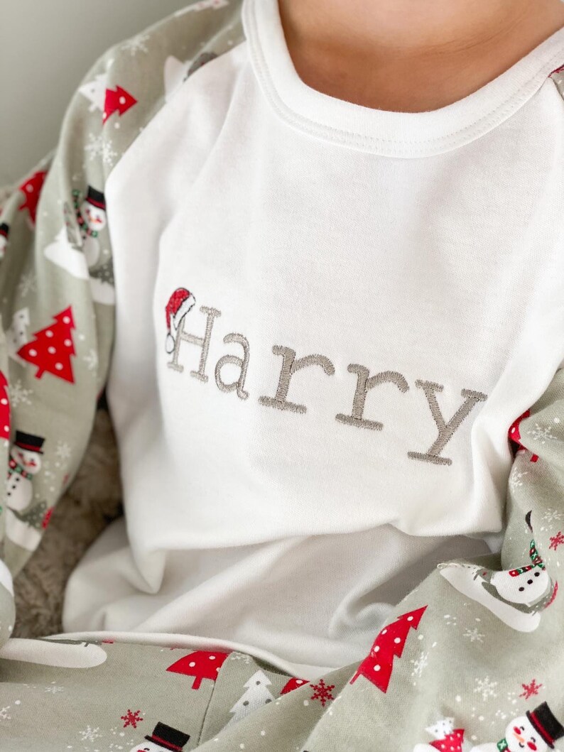 May include: White long-sleeve pajamas with a green and red snowman pattern. The top has a white collar and a gray embroidered name "Harry" with a red Santa hat.