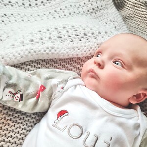 May include: A white baby onesie with a red Santa hat and the name "Louis" embroidered in gray. The baby is lying on a white crocheted blanket.