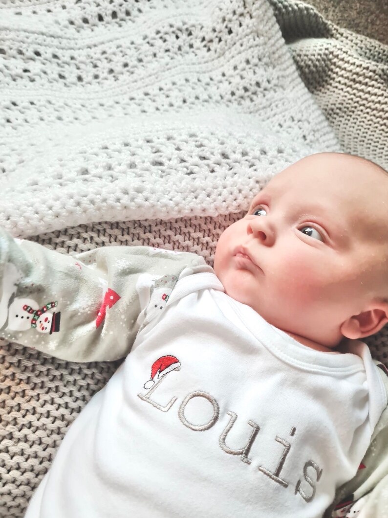 May include: A white baby onesie with a red Santa hat and the name "Louis" embroidered in gray. The baby is lying on a white crocheted blanket.