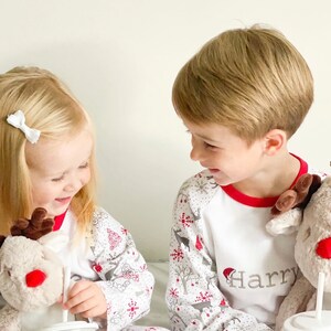 May include: Two children wearing white and red Christmas pajamas with reindeer and snowflake patterns. The boy's pajamas have the name "Harry" embroidered on the front. The girl's pajamas have the name "Belle" embroidered on the front. Both children are holding cups with straws.