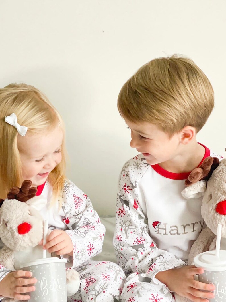 May include: Two children wearing white and red Christmas pajamas with reindeer and snowflake patterns. The boy's pajamas have the name "Harry" embroidered on the front. The girl's pajamas have the name "Belle" embroidered on the front. Both children are holding cups with straws.