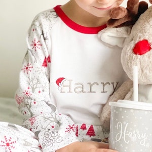 May include: A child wearing white pajamas with a red trim and a snowflake and reindeer print. The pajamas are personalized with the name "Harry". The child is holding a gray cup with a straw and the name "Harry" written on it.