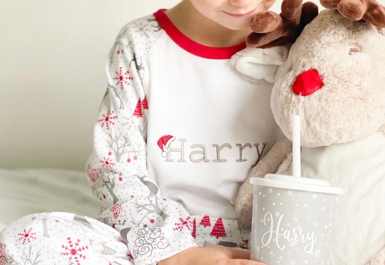 May include: A child wearing white pajamas with a red trim and a snowflake and reindeer print. The pajamas are personalized with the name "Harry". The child is holding a gray cup with a straw and the name "Harry" written on it.