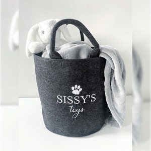 Könnte beinhalten: Ein grauer Stoffaufbewahrungskorb mit einem weißen Pfotenabdruck und dem Text "Sissy's toys" auf der Vorderseite. Der Korb ist gefüllt mit einem weißen Plüschkaninchen und einer grauen Decke.