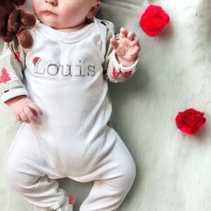 May include: A white baby onesie with long sleeves and a festive Christmas tree design. The onesie has the name "Louis" embroidered on the front in black thread.