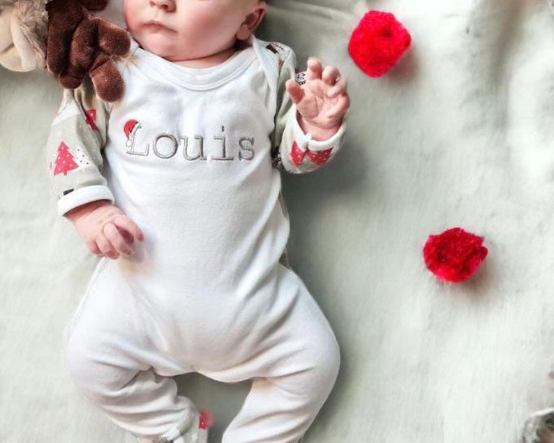 May include: A white baby onesie with long sleeves and a festive Christmas tree design. The onesie has the name "Louis" embroidered on the front in black thread.