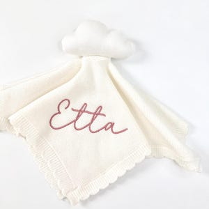 May include: A cream-coloured knitted security blanket featuring a plush cloud topper and a scalloped edge. The name Etta is embroidered in a dusty rose cursive script across the centre of the square fabric piece.