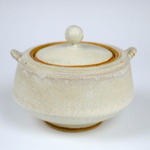 May include: A white ceramic covered dish with two handles and a knob on the lid. The dish has a speckled glaze and a brown rim.