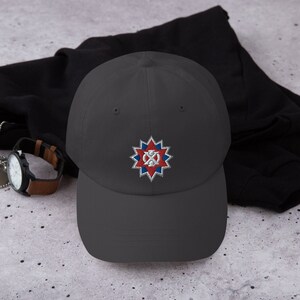 May include: A dark gray baseball cap with an embroidered patch featuring a red, white, and blue starburst design. The starburst has a white circle in the center with a red cross inside.