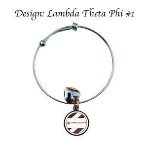 Könnte beinhalten: Ein silbernes, ausziehbares Armband mit einem Anhänger, der einen weißen und braunen Kreis mit dem Text "Lambda Theta Phi Latin Fraternity, Inc." zeigt. Der Anhänger ist an einem silbernen Herzen befestigt.