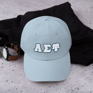Könnte beinhalten: Eine hellblaue Baseballkappe mit einem gestickten griechischen Buchstaben-Design in Weiß und Blau, das "AΣT" sagt.