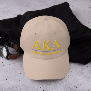 May include: A beige baseball cap with an embroidered gold "AKA" logo and the words "Alpha Kappa Lambda" in gold lettering.