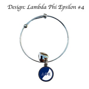 May include: A silver expandable bracelet with a charm featuring a blue and white circle with the letters "ΛΦΕ" in white. The charm is attached to a silver heart.