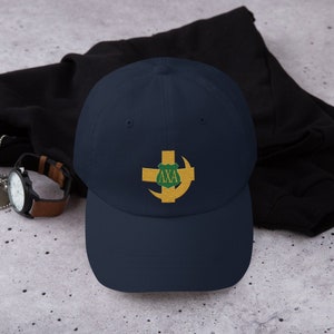 Può includere: Un cappello da baseball blu navy con una mezzaluna e una croce gialle ricamate con uno scudo verde e le lettere "AXA" in oro.