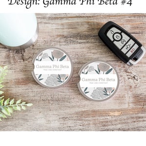 May include: Two round, gray car coasters with a floral design and the text "Gamma Phi Beta TRUE AND CONSTANT".