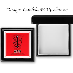 Könnte beinhalten: Eine schwarze Holzkiste mit einem roten und weißen Design. Die Kiste hat einen Klappdeckel und ein weißes Innenleben. Das Design zeigt einen Schild mit einem Wappen und den Buchstaben "ΑΠΥ" in Weiß. Der Text "Lambda Pi Upsilon Sorority, Latinas Poderosas Unidas, Inc." ist unter dem Schild gedruckt.