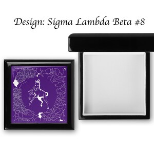 Könnte beinhalten: Eine schwarze Holzkiste mit einem Scharnierdeckel. Die Kiste ist geschlossen und der Deckel ist geöffnet. Das Innere der Kiste ist mit weißem Filz ausgekleidet. Die Oberseite der Kiste hat einen violetten Hintergrund mit einer weißen Silhouette eines Pferdes und einem weißen Design. Der Text "Design: Sigma Lambda Beta #8" ist auf der Oberseite der Kiste aufgedruckt.