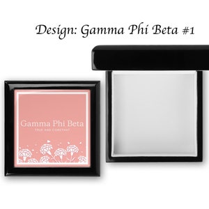 May include: A black wooden box with a hinged lid. The lid is open and the inside is lined with white felt. The front of the box has a pink background with white text that reads "Gamma Phi Beta True and Constant" and a white floral design.