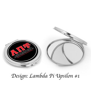 Op de afbeelding: Een zilveren compact spiegel met een zwarte achtergrond en rode letters die "ΑΠΥ LAMBDA PI UPSILON SORORITY. LATINAS PODEROSAS UNIDAS, INC." lezen.