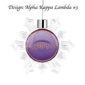 May include: A purple and gold snowflake ornament with the letters "AKA" inside a stylized eye. The ornament is hanging from a silver string.