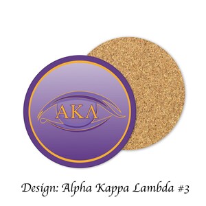 May include: Purple and gold round coaster with an eye design and the letters "AKA" inside. The coaster is made of cork.