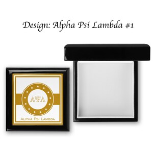 Op de afbeelding: Een zwarte houten doos met een gouden ontwerp met de letters "ΑΨΛ" en de tekst "Alpha Psi Lambda". De doos is open en de binnenkant is bekleed met wit vilt.